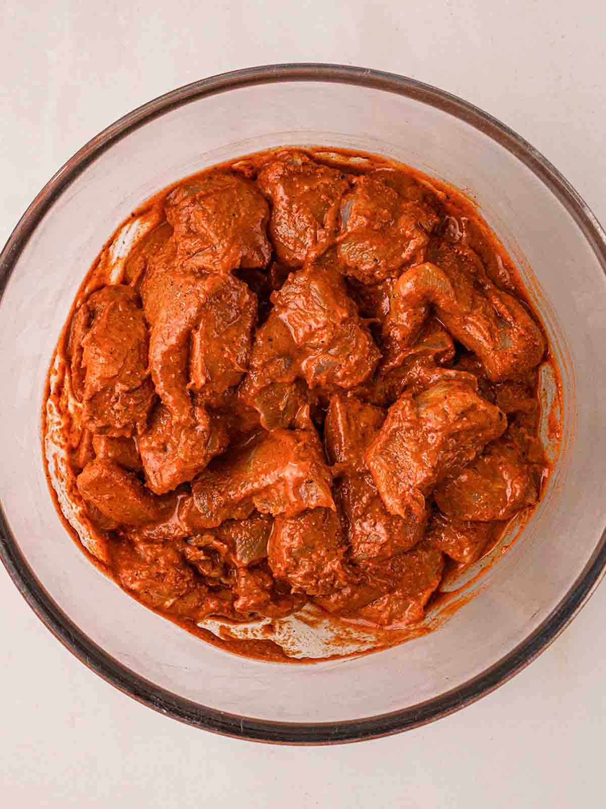 A glass bowl filled with chunks of raw chicken in a tandoori marinade for step 1 in the recipe.